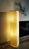 A tall, rectangular floor lamp with a textured beige paper shade is illuminated in a room, casting a warm glow on the wall. Decorative metal flowers are mounted on the wall behind the lamp. Produktbild-Vorschau