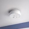 A smoke detector is mounted on a white ceiling, with a blue stripe visible on the wall below. Produktbild-Vorschau