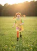 A young girl with red pigtails runs through a grassy field at sunset, wearing a yellow dress over a green shirt and colorful tights. Produktbild-Vorschau
