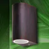 A dark brown, modern outdoor wall light with a curved design is mounted on a textured green background. Produktbild-Vorschau