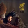 A young girl lies in bed, illuminated by the light of a tablet, with a large teddy bear beside her and a unicorn lamp glowing on a shelf. Produktbild-Vorschau