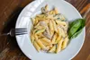 A close-up shot of a plate of penne pasta with mushrooms and spinach, garnished with parmesan cheese and a basil leaf, on a wooden table. Produktbild-Vorschau