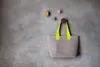 A woven tote bag with yellow handles hangs from a hook on a textured gray wall. Several colorful circular hooks are also visible on the wall. Produktbild-Vorschau