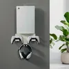 A white Playstation 5 console, two controllers, and headphones are mounted on a gray wall. A potted plant is visible on the right side of the image. Náhľad obrázku produktu