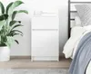 A white bedside table with two drawers is shown next to a bed and a large potted plant. Podgląd zdjęcia produktu