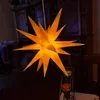 A yellow star-shaped lamp is hanging in a dimly lit room, with a wooden cabinet in the background and a candle holder on a table in the foreground. Produktbild-Vorschau