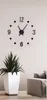 A wall clock with black numbers and hands is mounted on a light-colored wall. In the foreground, a white planter with a green plant sits on a wooden floor, and a portion of a gray sofa is visible on the right. Náhled obrázku produktu