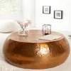 A round copper coffee table with a hammered finish is shown in a living room setting, with a white pouf and a vase of flowers.