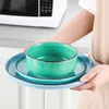 A person holding a stack of turquoise and blue ceramic plates and bowls in front of a microwave. Produktbild-Vorschau