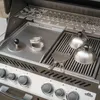 A close-up view of a stainless steel grill with various accessories, including a burger press, burger domes, and a spatula. Podgląd zdjęcia produktu
