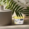 A small container of air freshener sits on a surface next to a larger gray object, with green leaves in the background. Anteprima dell’immagine prodotto