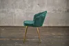 A green velvet chair with gold legs is shown against a concrete wall and wooden floor. Produktbild-Vorschau