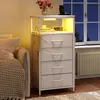 A white nightstand with drawers and shelves, illuminated by LED lights, is placed next to a bed in a bedroom setting. A vase of pink flowers and a wireless charger are on top of the nightstand. A window with a view of the night sky is visible in the backgr Produktbild-Vorschau