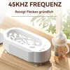 A white ultrasonic cleaner is shown on a wooden surface next to a baby bottle, with plants in the background. Produktbild-Vorschau