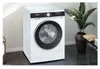 A white front-loading washing machine is shown in a modern laundry room setting, next to a large potted plant and a shelf with folded towels. The floor is dark gray. Produktbild-Vorschau