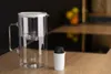 A glass water filter pitcher and a replacement filter cartridge are placed on a wooden countertop in a kitchen. Produktbild-Vorschau