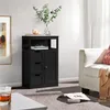 A black wooden cabinet with drawers and a door, placed in a living room setting next to a white armchair. Produktbild-Vorschau