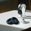 A pair of black ear pads with blue inner lining sits on a white surface next to a white coffee cup and saucer. A pair of black headphones rests on a white stand in the background. Produktbild-Vorschau