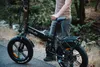 A person stands next to a black electric bicycle on a paved path with trees and a stream in the background. Produktbild-Vorschau