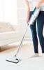 A person is vacuuming a carpeted floor with a cordless stick vacuum cleaner in a bright, modern living room. Náhled obrázku produktu