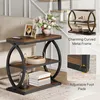 A narrow, three-tier side table with a rustic brown wood finish and black metal frame, placed next to a sofa. The table is decorated with a vase of flowers, framed pictures, and books. Anteprima dell’immagine prodotto