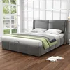 A bedroom scene with a grey upholstered bed, dressed with white bedding, a green throw blanket, and a grey and white patterned blanket. Produktbild-Vorschau