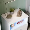 A white nightstand with a lamp, plant, and clock on top, and books and a cube on the shelf. Produktbild-Vorschau