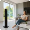 A woman is reading a book on a couch while a tower fan operates in the background. Náhled obrázku produktu