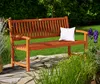 A wooden garden bench with a white cushion sits on a stone patio, surrounded by lush greenery and purple flowers. Náhled obrázku produktu