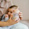 A person is holding a baby bottle with milk in it, with a baby in the background. Anteprima dell’immagine prodotto