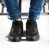 A close-up view of a person's legs and feet wearing black sneakers and jeans, with the feet resting on a metallic platform. Náhľad obrázku produktu