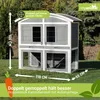 A two-story rabbit hutch with an outdoor run, set on a grassy lawn with trees in the background. Dimensions and features are highlighted with text overlays and graphics. Produktbild-Vorschau
