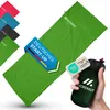 A green sleeping bag is laid out diagonally, with several other colored sleeping bags visible in the background. A hand holds a small, dark green pouch with a tag attached. Promotional graphics are overlaid on the sleeping bag. Produktbild-Vorschau