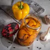 A still life arrangement of preserved bell peppers in a jar, a whole yellow bell pepper, a jar lid with a fruit pattern, and garlic bulbs on a light gray surface with a textured gray cloth. Podgląd zdjęcia produktu
