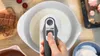 A hand is using a white and gray hand mixer to whip cream in a white bowl. Chocolate bars and cherries are visible in the background. Produktbild-Vorschau