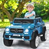 A young boy is happily driving a blue toy Mercedes-Benz G-Wagon in an outdoor setting with trees and greenery.