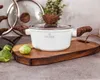 A white pot with wooden handles and lid sits on a wooden cutting board, with Brussels sprouts in the foreground. Produktbild-Vorschau