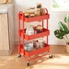 A red kitchen cart filled with jars of food items and bowls, placed on a wooden floor next to a kitchen counter and a plant. Produktbild-Vorschau