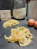 A close-up shot of freshly made pasta on a dark gray surface, with ingredients like flour, eggs, and a box labeled 'PASTA DISC' in the background. Voir les images du produit