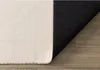 The underside of a cream-colored rug with a black anti-slip backing is shown on a wooden floor. Produktbild-Vorschau
