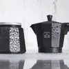 Two black coffee makers are on a reflective surface, covered in water droplets, against a textured white background. Produktbild-Vorschau