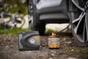 A car tire inflator and sealant kit is shown next to a car tire on a gravel surface, with a bicycle in the background. Produktbild-Vorschau