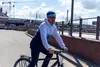 A man is riding a bicycle on a paved path with an urban background. Náhľad obrázku produktu
