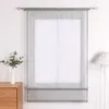 A sheer Roman blind hangs in front of a window, with a vase of greenery on a wooden dresser to the left and a pleated lamp to the right. Produktbild-Vorschau