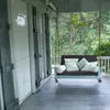 A porch swing with cushions on a wooden porch, with trees in the background. Podgląd zdjęcia produktu