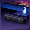A black soundbar with blue LED lights is placed on a dark wooden surface in front of a computer monitor. The monitor displays a colorful image and is illuminated by blue ambient lighting. Náhled obrázku produktu