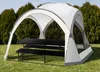 A white and gray outdoor canopy tent with black picnic tables and benches set up on a grassy lawn, with trees in the background. Podgląd zdjęcia produktu