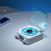A white, vintage-style speaker with a clear blue lid, resembling a record player, sits on a white surface. A book is visible to the left, and a blue background is behind. Podgląd zdjęcia produktu