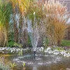 A solar-powered fountain kit is shown operating in a pond surrounded by tall grasses and rocks. Produktbild-Vorschau