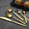 A set of gold-colored cutlery is arranged on a dark surface with a wire rack and a plate of food in the background. Anteprima dell’immagine prodotto